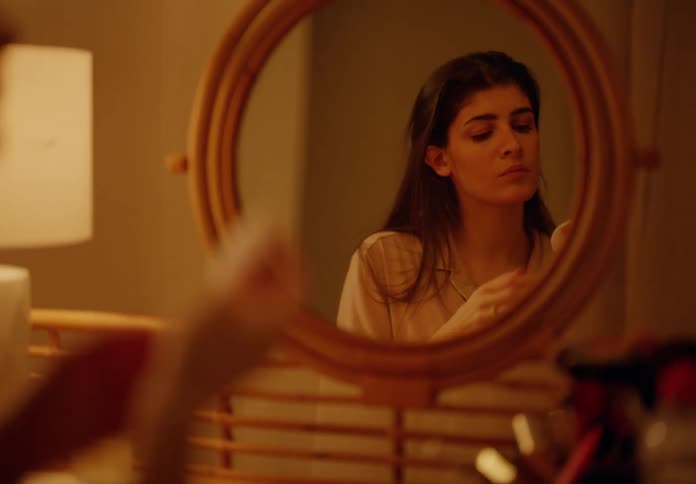 A woman is doing skin care on her face while looking at herself in a mirror with orange ambient light