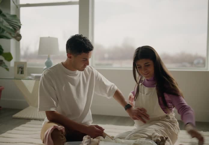 A man and a woman are sitting on the floor of a room and they are looking at clothes that are on the floor and the man is grabbing the womans belly