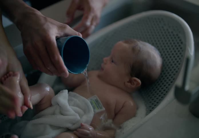 Two adult hands gently pouring water over a baby inside a bathtub