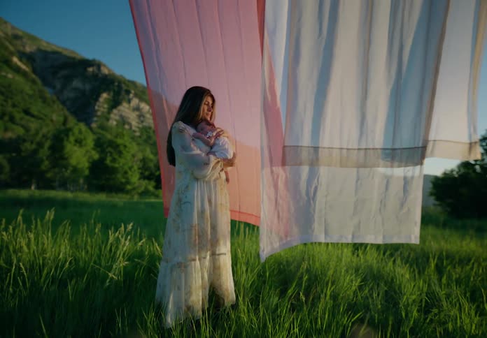 A mother standing in a grassy field holding a baby with clothes hung on a line