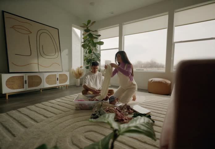 A man and a pregnant woman are sitting on the carpet of a living room floor and fiddling with baby clothes