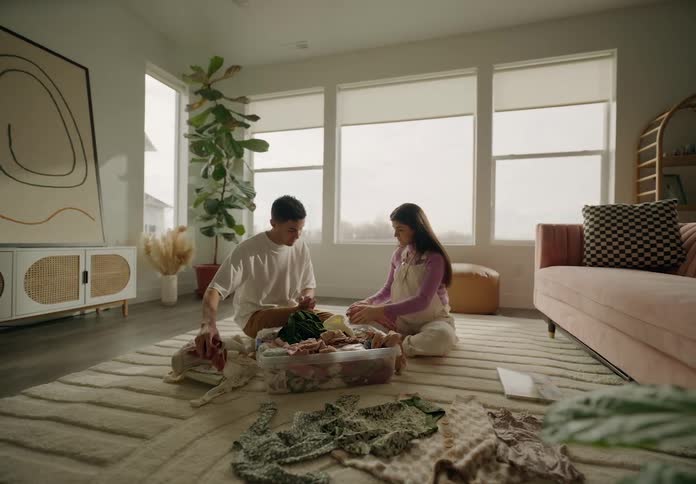 A man and a woman are sitting on the floor of a living room and are fiddling with baby clothes that are in a plastic basin