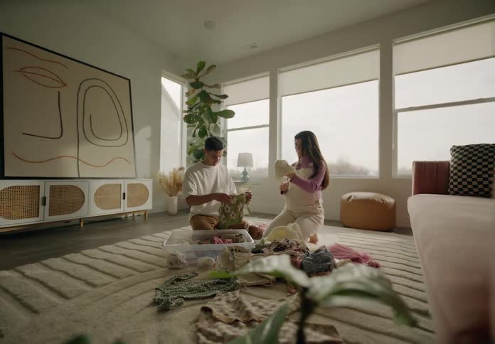 A couple sitting on the floor folding laundry from a basket in a welllit cozy living room with plants and furniture
