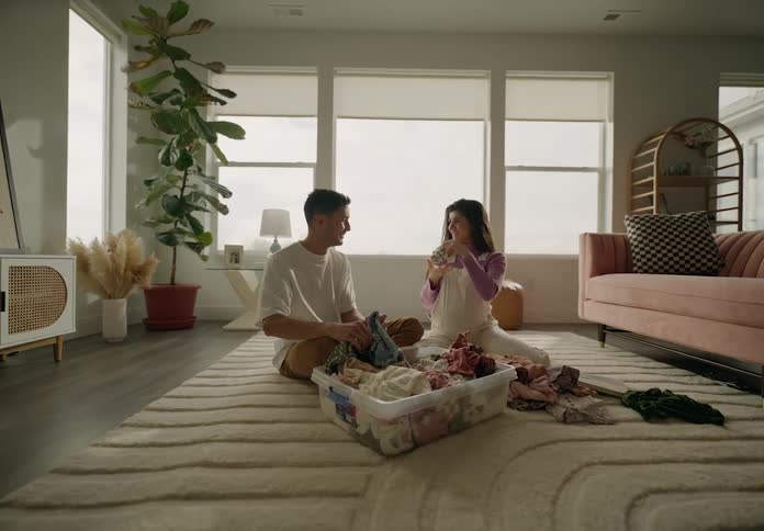 A couple sitting on the floor folding laundry from a basket in a welllit cozy living room with plants and furniture