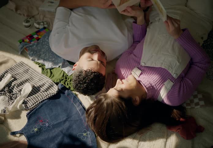 A couple lies on the bed their heads resting against each other reading a book in the morning