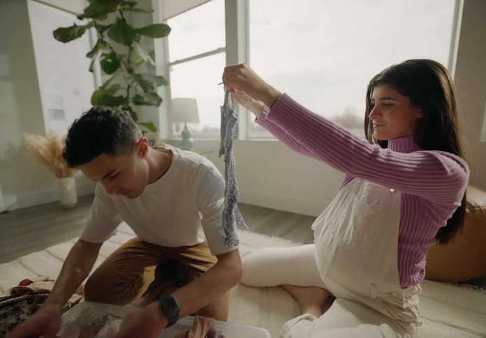 Pregnant woman looking at baby clothes next to her husband in the living room sitting on the carpet