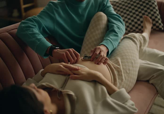 A man plays a music box on a pregnant womans belly as she rests on the couch at home
