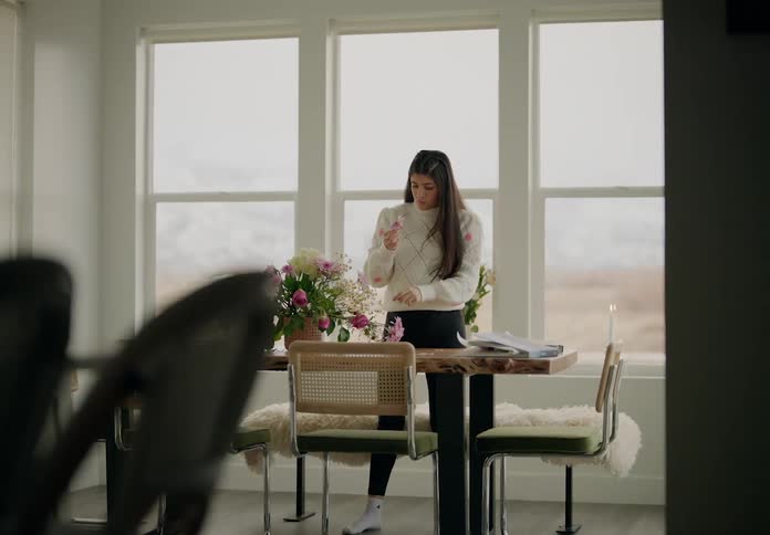 A woman stands next to large windows arranging a bouquet of pink and white flowers