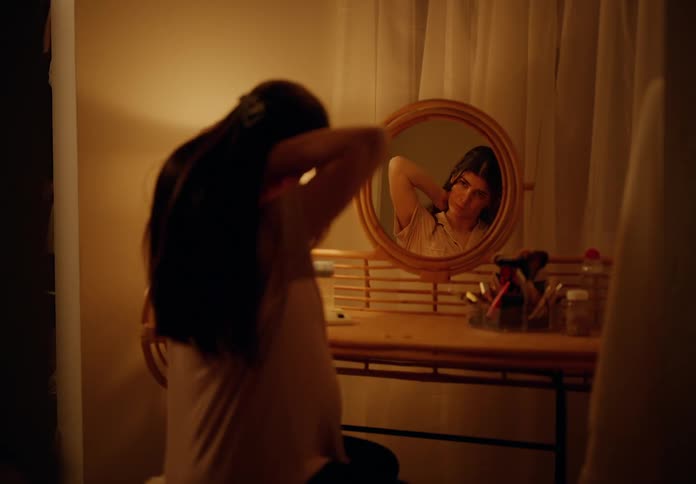 A woman sitting in front of the bedside table arranging earrings and a necklace in front of the mirror