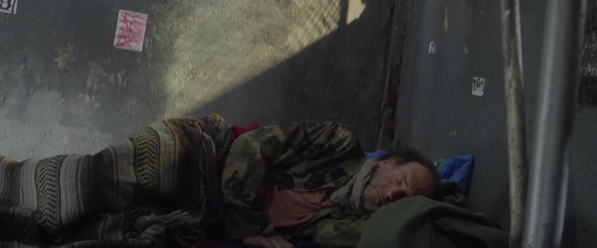 A homeless man lies down against a wall to sleep