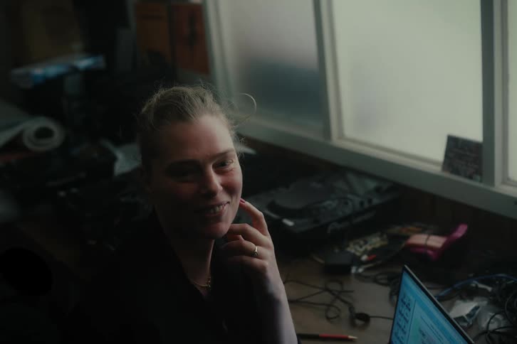 A woman is smiling and putting her hand on her cheek and in front of her there is a table full of things and a laptop