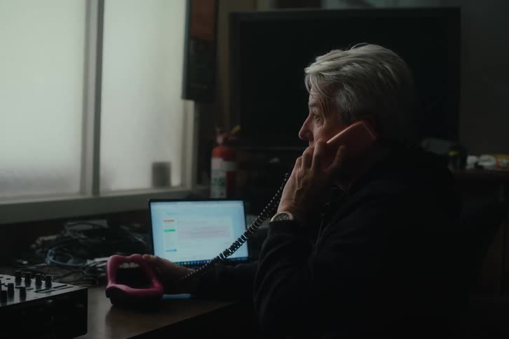 An elderly man sitting inside a dimly lit broadcasting studio talking on a vintage novelty phone