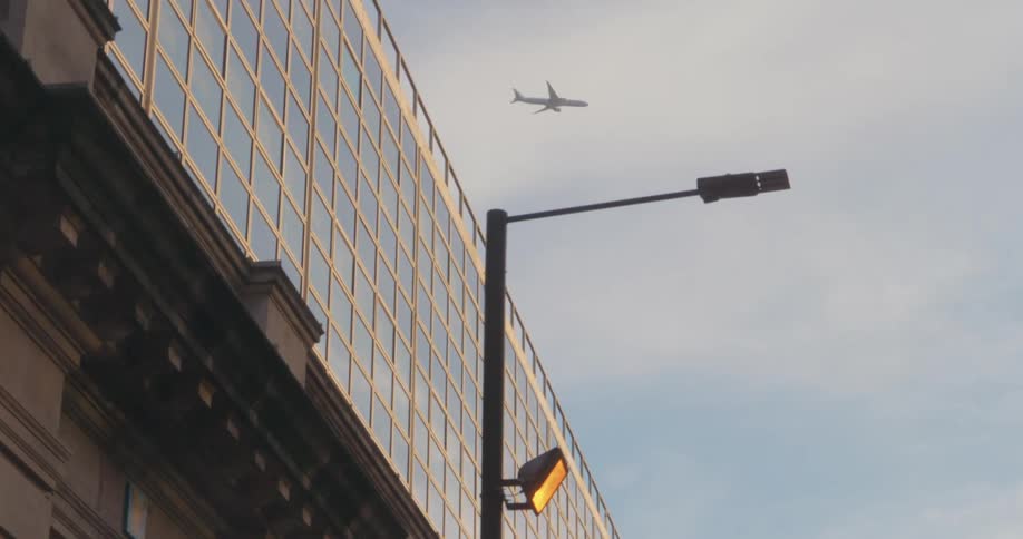 A plane flying above a cityscape with a modern glass building and a streetlight in the foreground