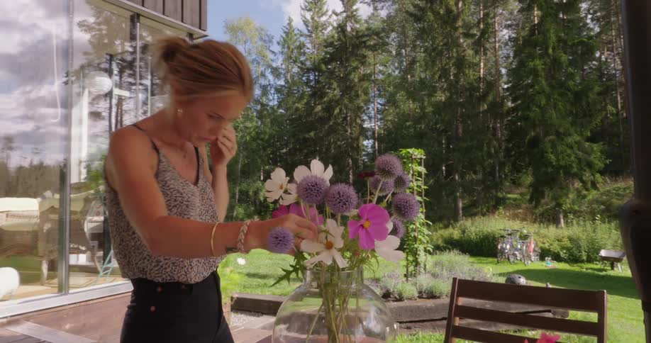 A blonde woman is arranging a flower in a glass vase that already has several of them