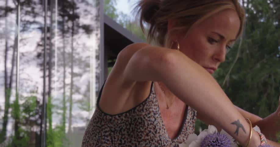 A woman arranges purple white and pink flowers in a clear glass vase outdoors