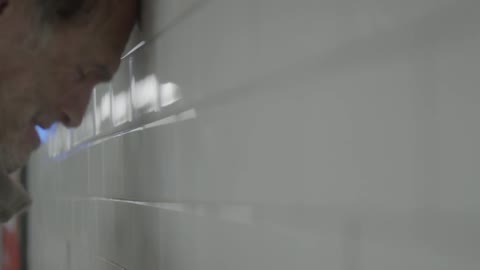 An old man leans his head against a tiled walled
