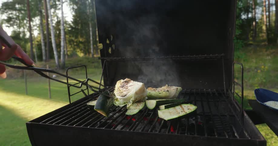 Vegetables including zucchini and cabbage grilling on a barbecue with smoke rising in a natural outdoor setting