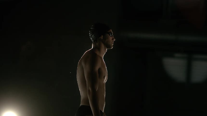 Male swimmer wearing swim goggles sheds water while standing in the dark