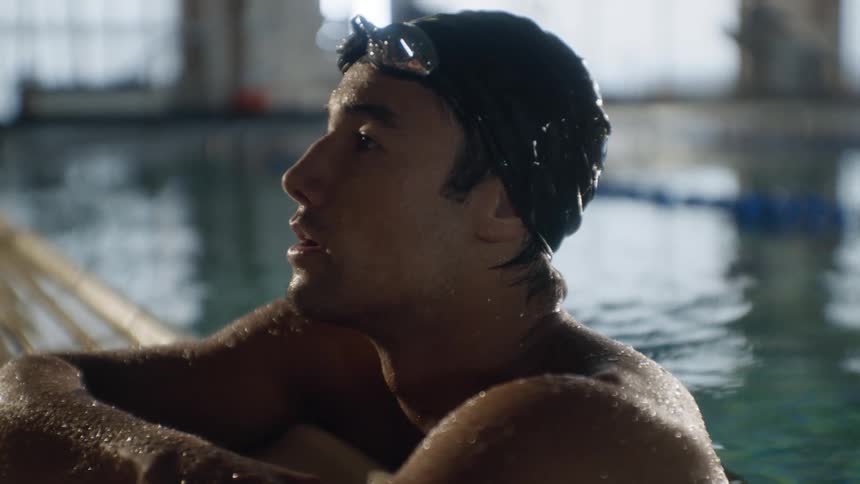 A male competitive swimmer inside a pool resting while leaning against the edge looking sad