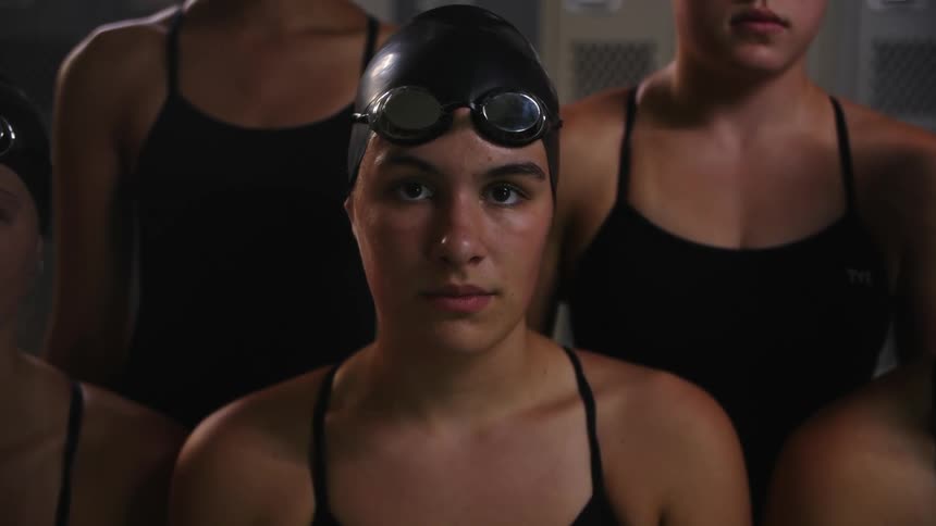 A woman dressed in black swimwear sits closely with teammates in front of a row of grey lockers