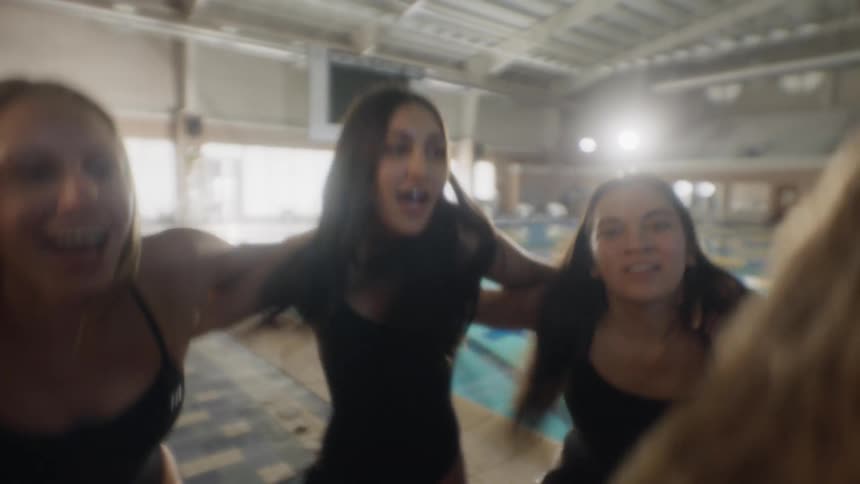 Group of women in swimsuits next to a swimming pool holding each other close raising their hands in celebration