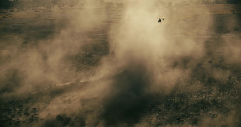 A helicopter flying over a landscape covered in dust raised by a running herd of buffaloes