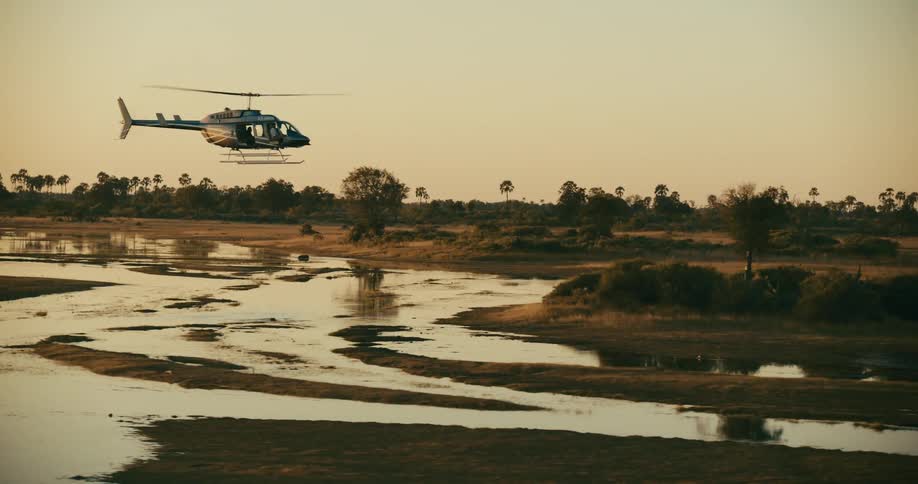 A helicopter flies over a shallow river that runs through a savanna