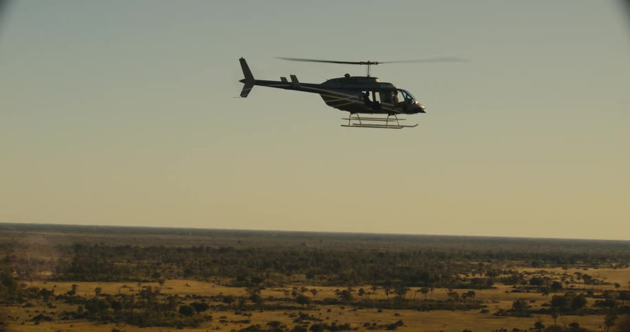 A helicopter flies over a partially forested grassy plain