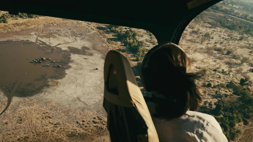 Cockpit view from a helicopter shows a pilot over a landscape with several elephants nearby a dry lake
