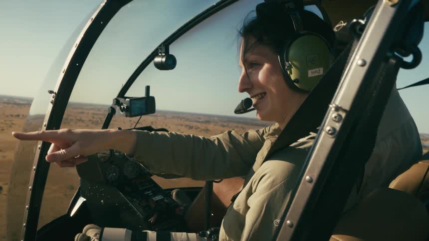 A woman in a helicopter smiling and pointing as it flies over a savanna