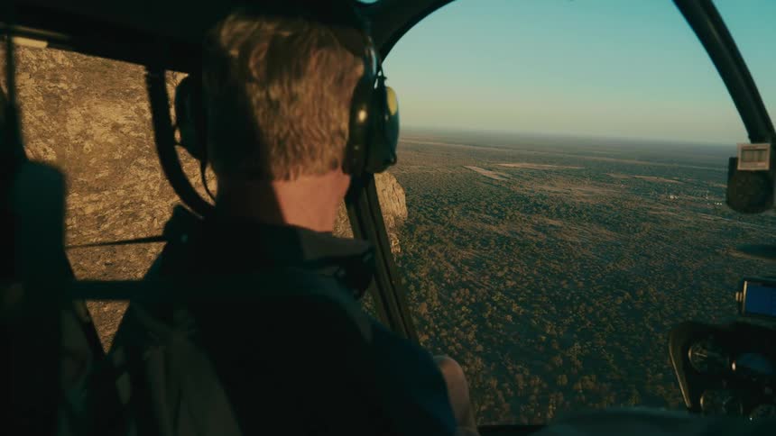 Two people fly in a helicopter next to a large mountain and over a vast savanna