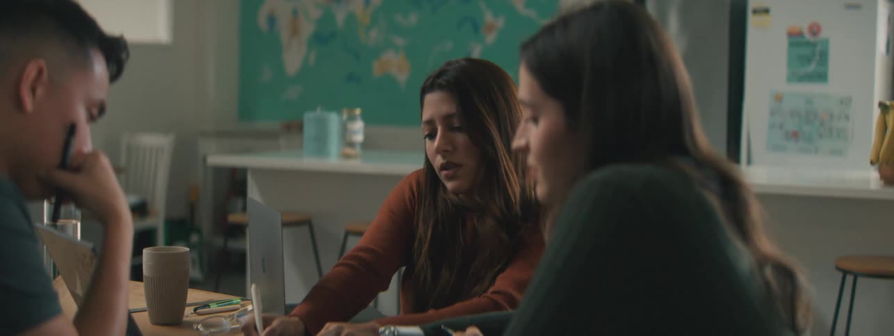 Two women and a man are around a table with laptops and notebooks and are talking while one woman writes in a notebook and the other woman with a mechanical hand points to the notebook