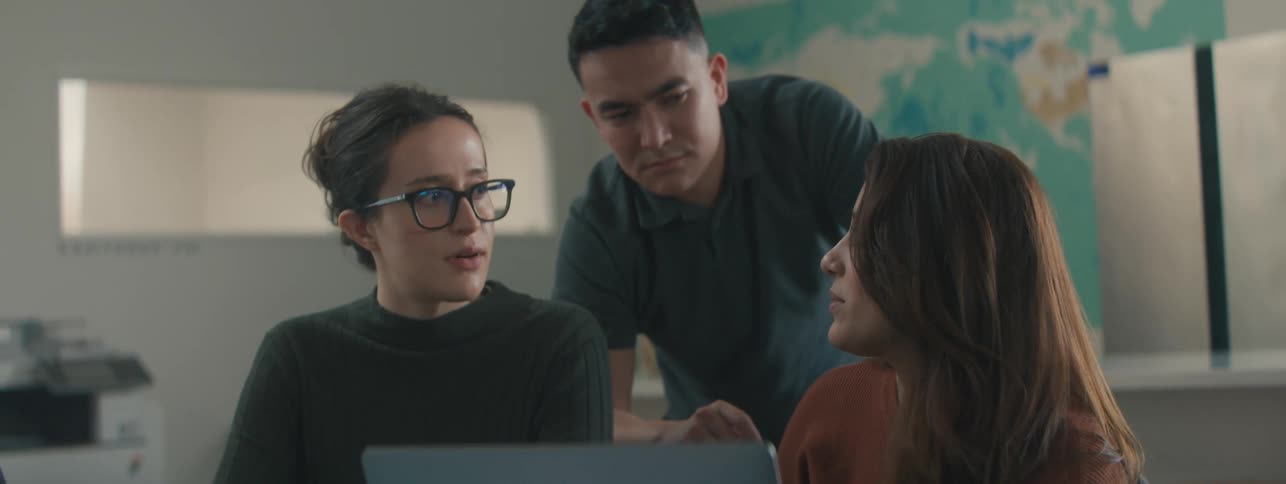Two women sitting and a man standing behind them as they talk with a laptop on the table in front of them