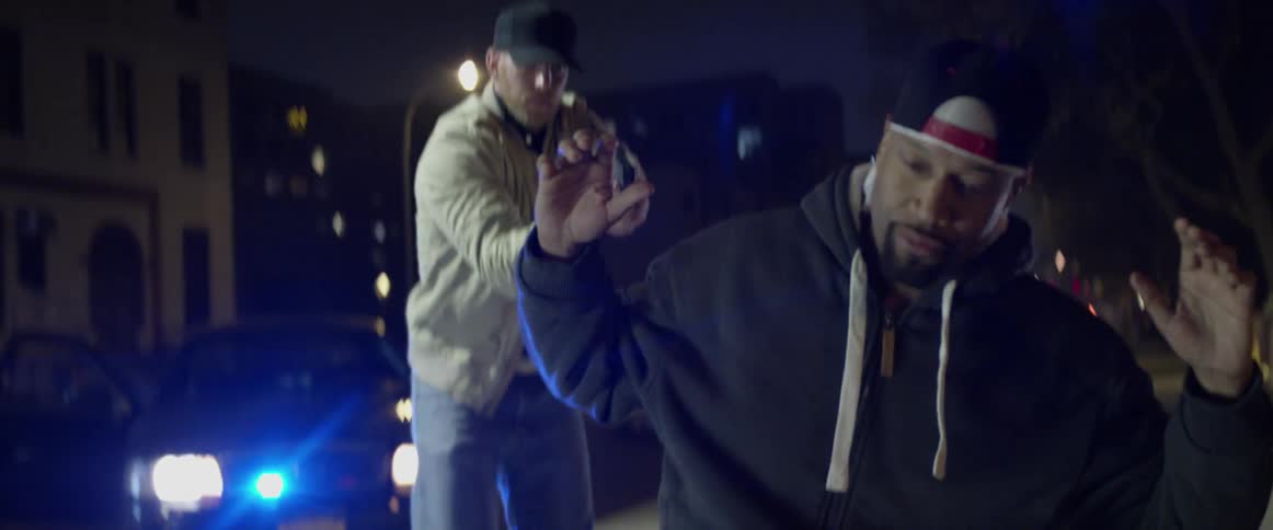 A black man kneels to the ground as a cop is pointing a gun at him