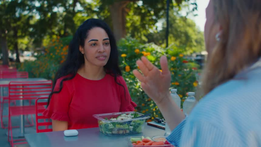 Two women speak while sitting at an outdoor restaurant preparing to eat