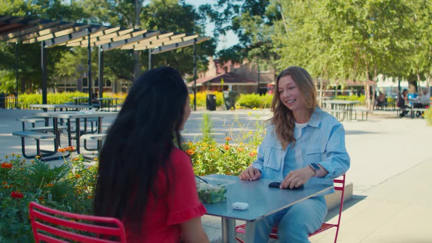 Woman joins a friend eating lunch at an outdoor table in the park on a sunny day