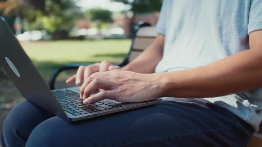 Person sitting on a bench in a square typing on a laptop