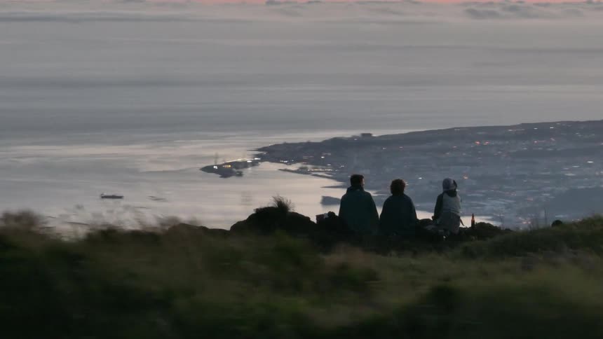 Three people sit on a promontory on a foggy day looking down onto a coastal town under ared tinted overcast sky