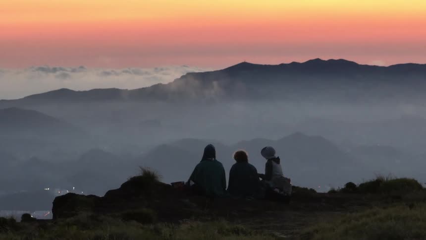 Three people are eating while sitting on a mountaintop at dusk