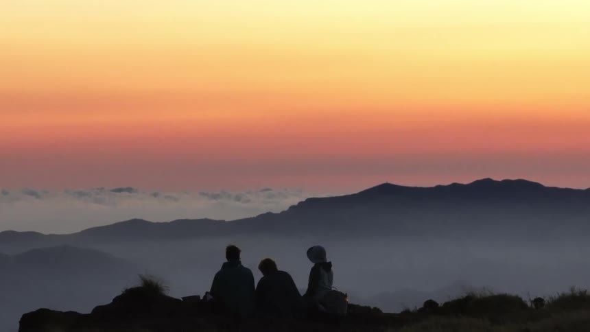 Three people sit on the edge of a mountain overlooking a small community nestled in a valley