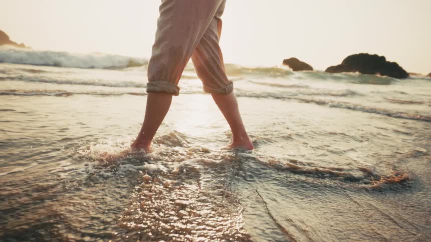 A woman with her pants rolled up strolls through the incoming surf at a beach