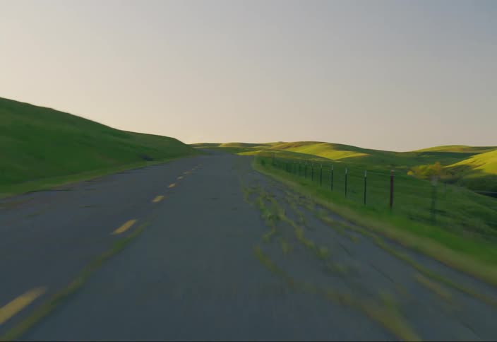 An asphalt road with yellow lines in the middle and on the right side some hills covered with green grass