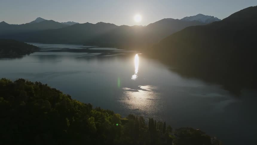 The sun hangs low over a mountain range while shining light on a lake