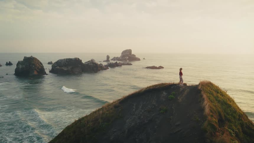A blonde woman stands on a mountaintop overlooking the ocean shore at dusk