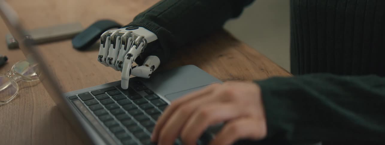Pair of hands one of which is robotic typing on a laptop
