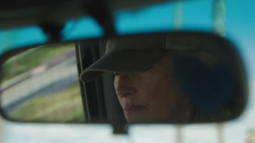 A persons reflection in a cars rearview mirror reveals a glimpse of their face and a baseball cap