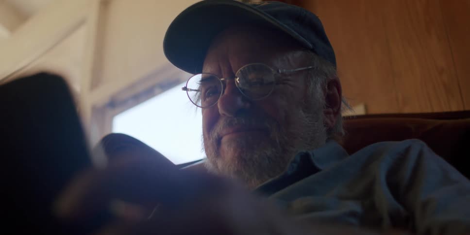 An elderly man puts on his glasses then looks down at the tablet hes holding and laughs
