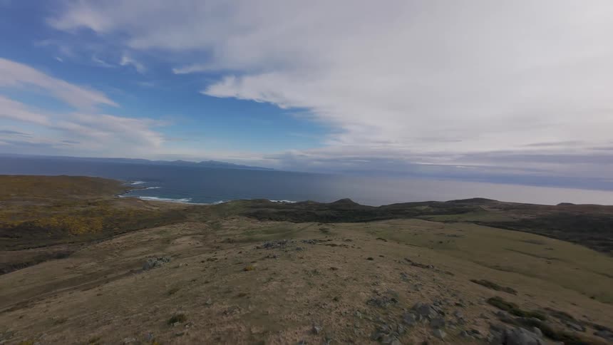 An aerial FPV view of a large field with hills and rocks and lots of low grass towards the rugged blue ocean