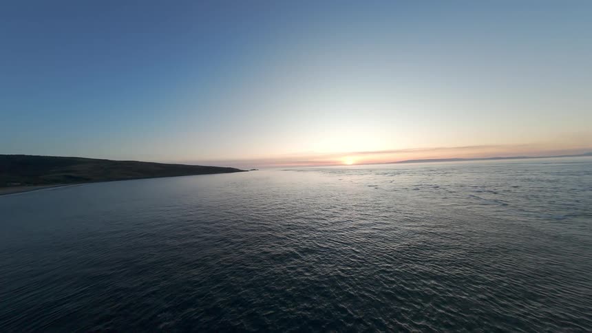 FPV drone across calm blue sea with the sun rising over the horizon in moody low light