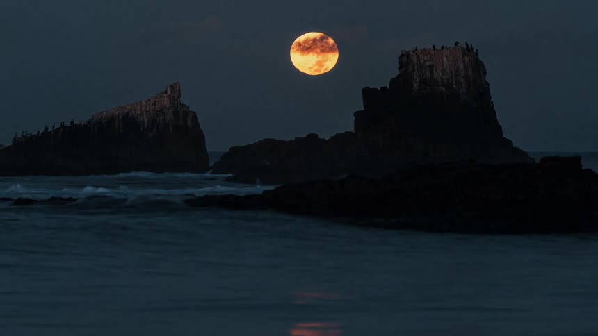 Moon on the horizon above the sea with rocks in the background